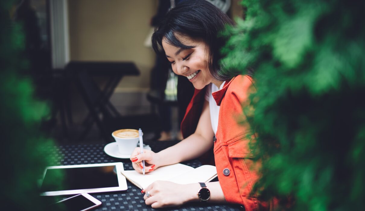 Excited ethnic woman writing down ideas in outdoor cafeteria