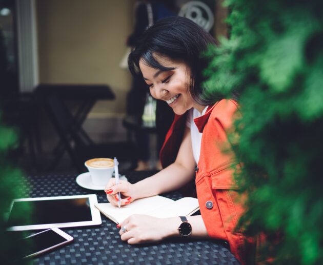 Excited ethnic woman writing down ideas in outdoor cafeteria