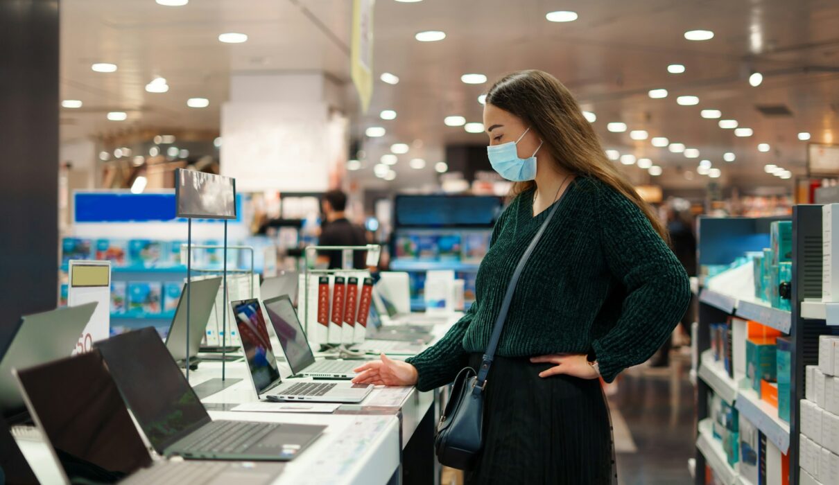 Focused young woman in face mask examines laptop on display in tech store department