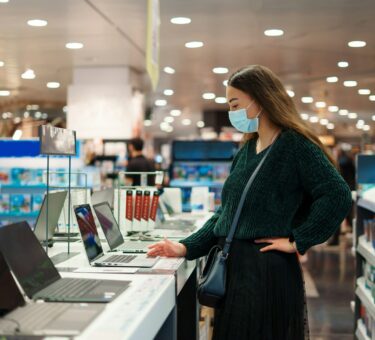 Focused young woman in face mask examines laptop on display in tech store department