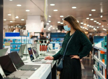 Focused young woman in face mask examines laptop on display in tech store department