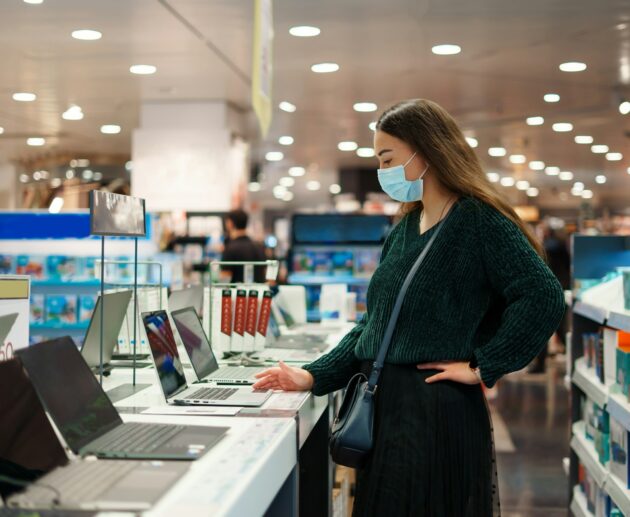 Focused young woman in face mask examines laptop on display in tech store department