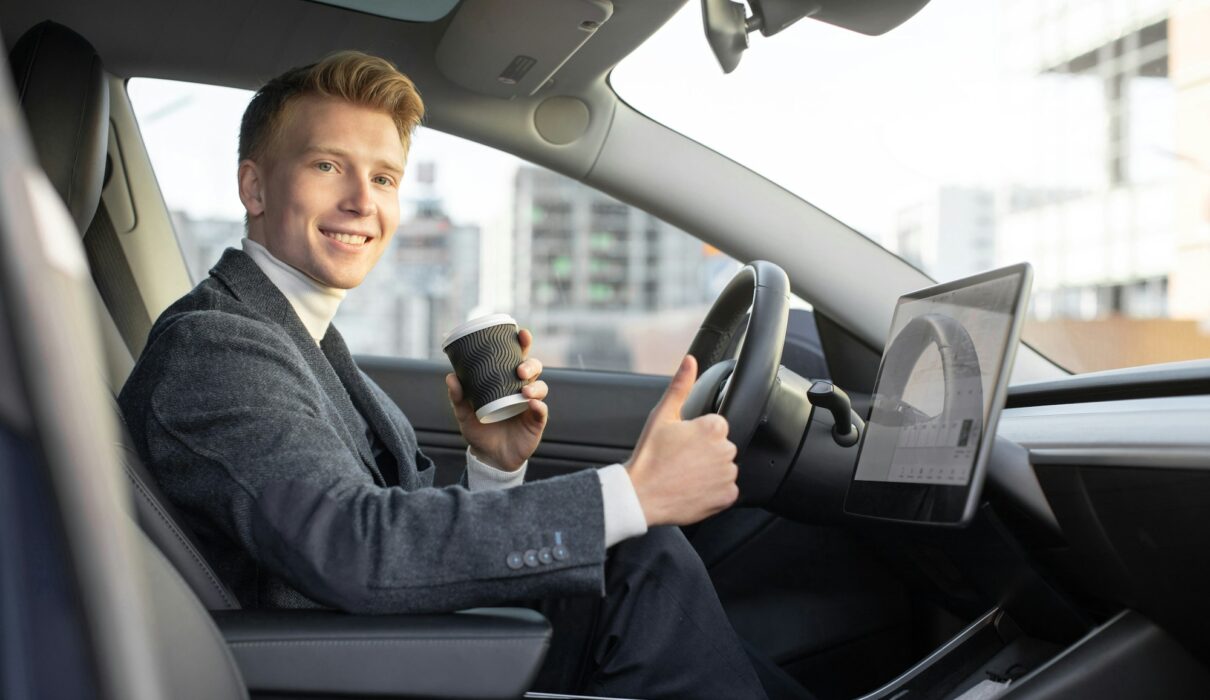 Handsome smiling Caucasian business man riding autonomous car, drinking coffee to go