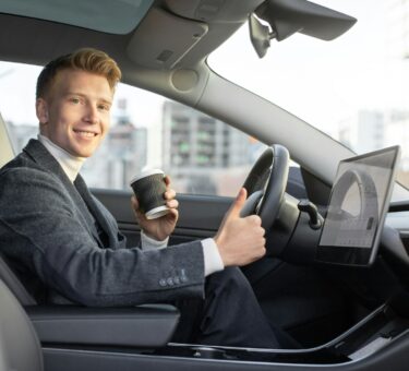 Handsome smiling Caucasian business man riding autonomous car, drinking coffee to go