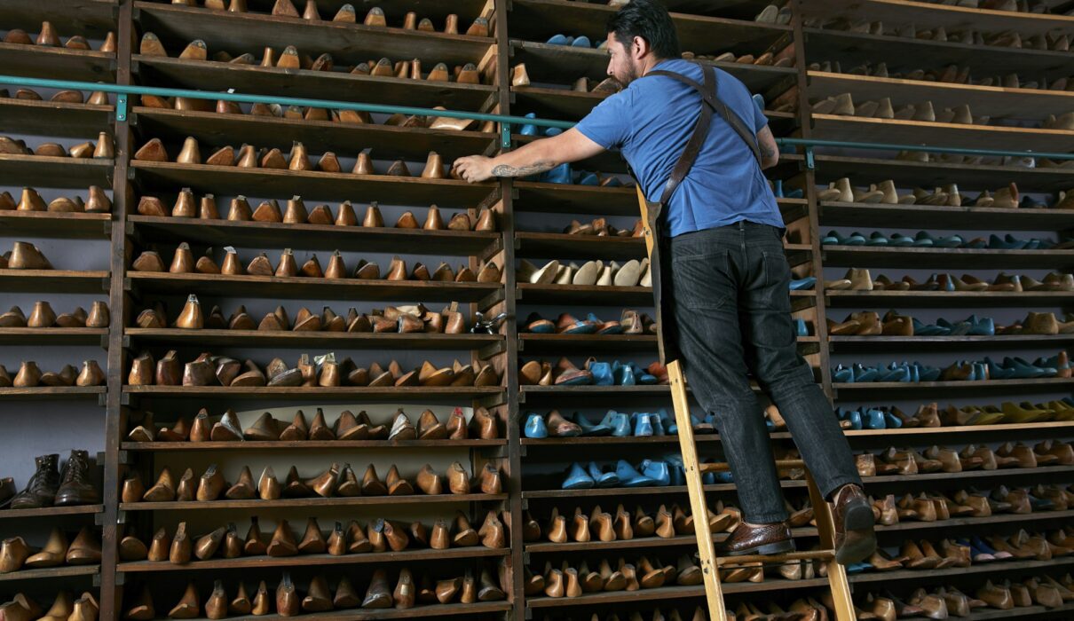 Male cobbler in traditional shoe shop on ladder selecting shoe last