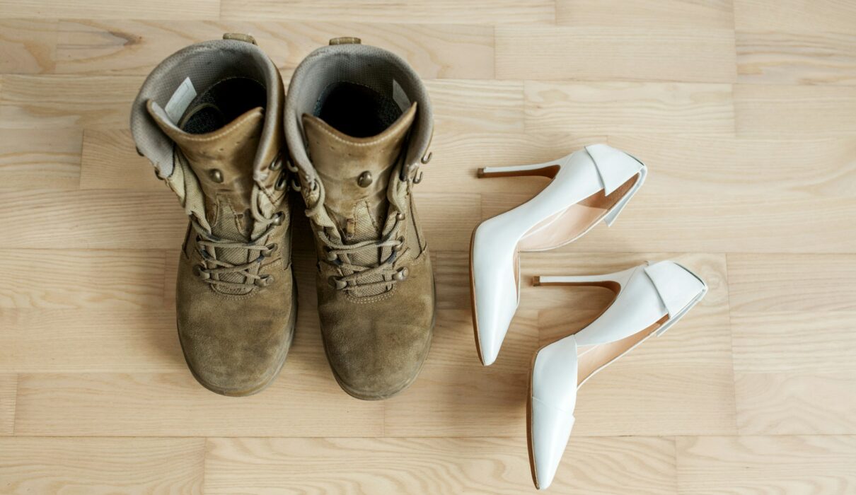 Old worn military boots, women's white shoes on wooden floor. Veteran and family concept.