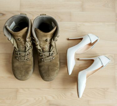 Old worn military boots, women's white shoes on wooden floor. Veteran and family concept.