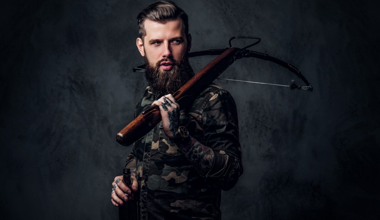 Stylish bearded hipster man in military shirt holding a craft beer and a medieval crossbow