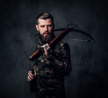 Stylish bearded hipster man in military shirt holding a craft beer and a medieval crossbow