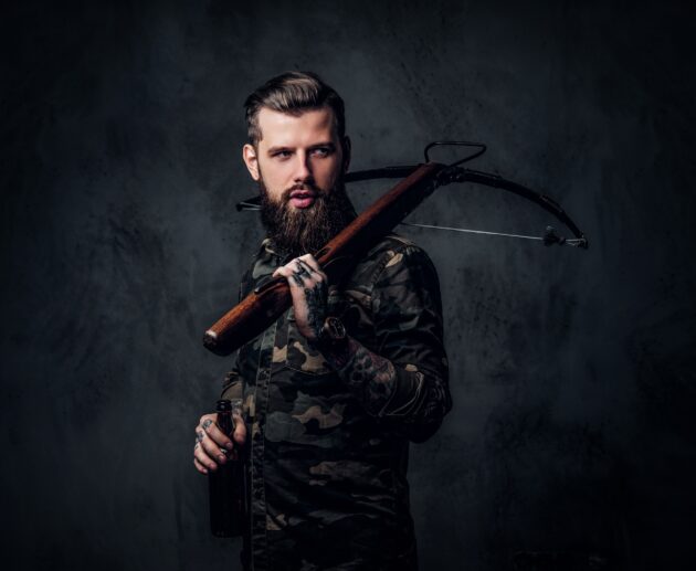 Stylish bearded hipster man in military shirt holding a craft beer and a medieval crossbow