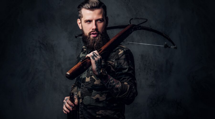 Stylish bearded hipster man in military shirt holding a craft beer and a medieval crossbow